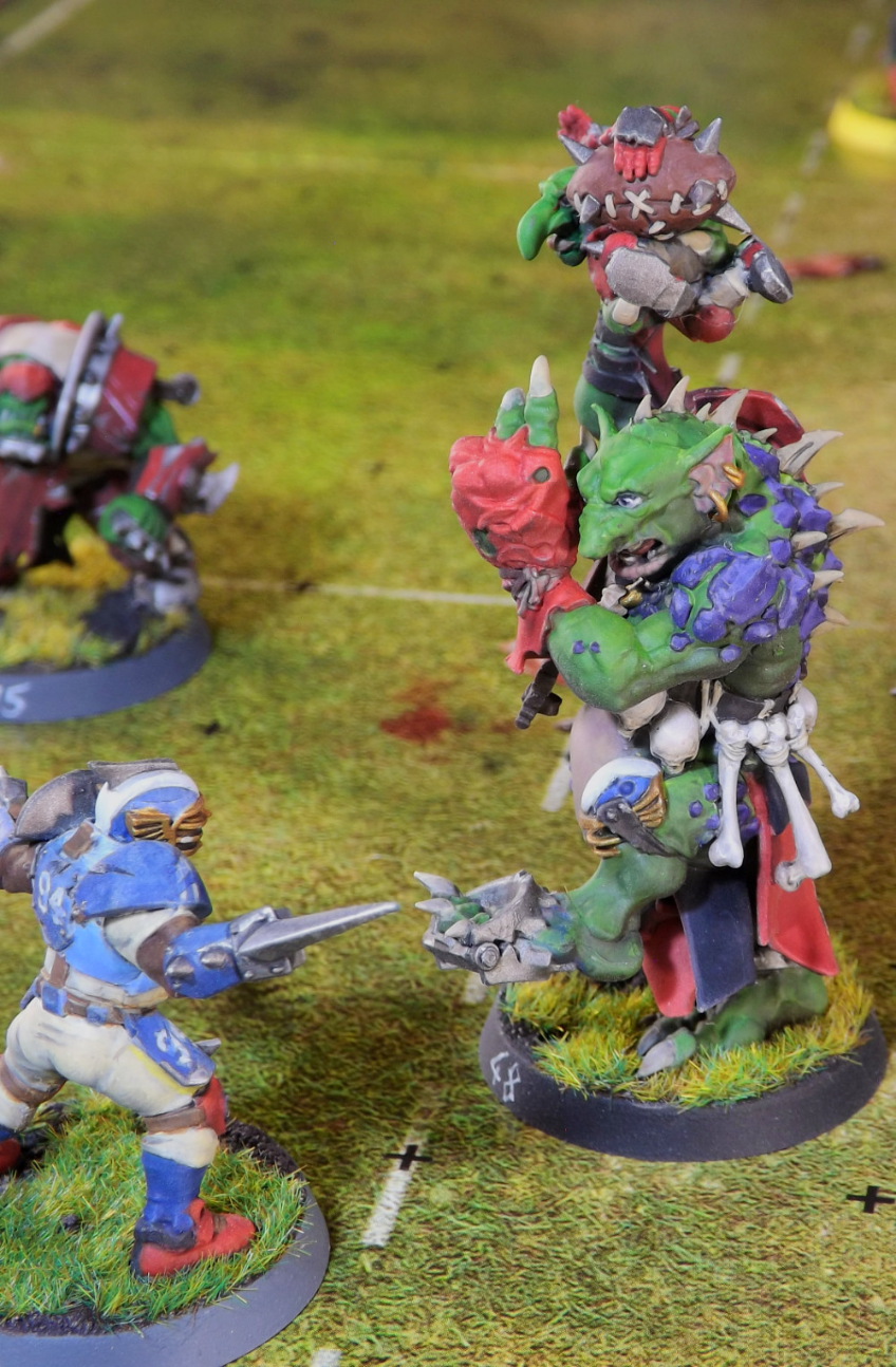 Photograph of an Orc Blood Bowl team playing against a Human Team on the table top. Miniatures are painted by Haximica. Green and purple painted blood bowl troll facing off a human lineman.